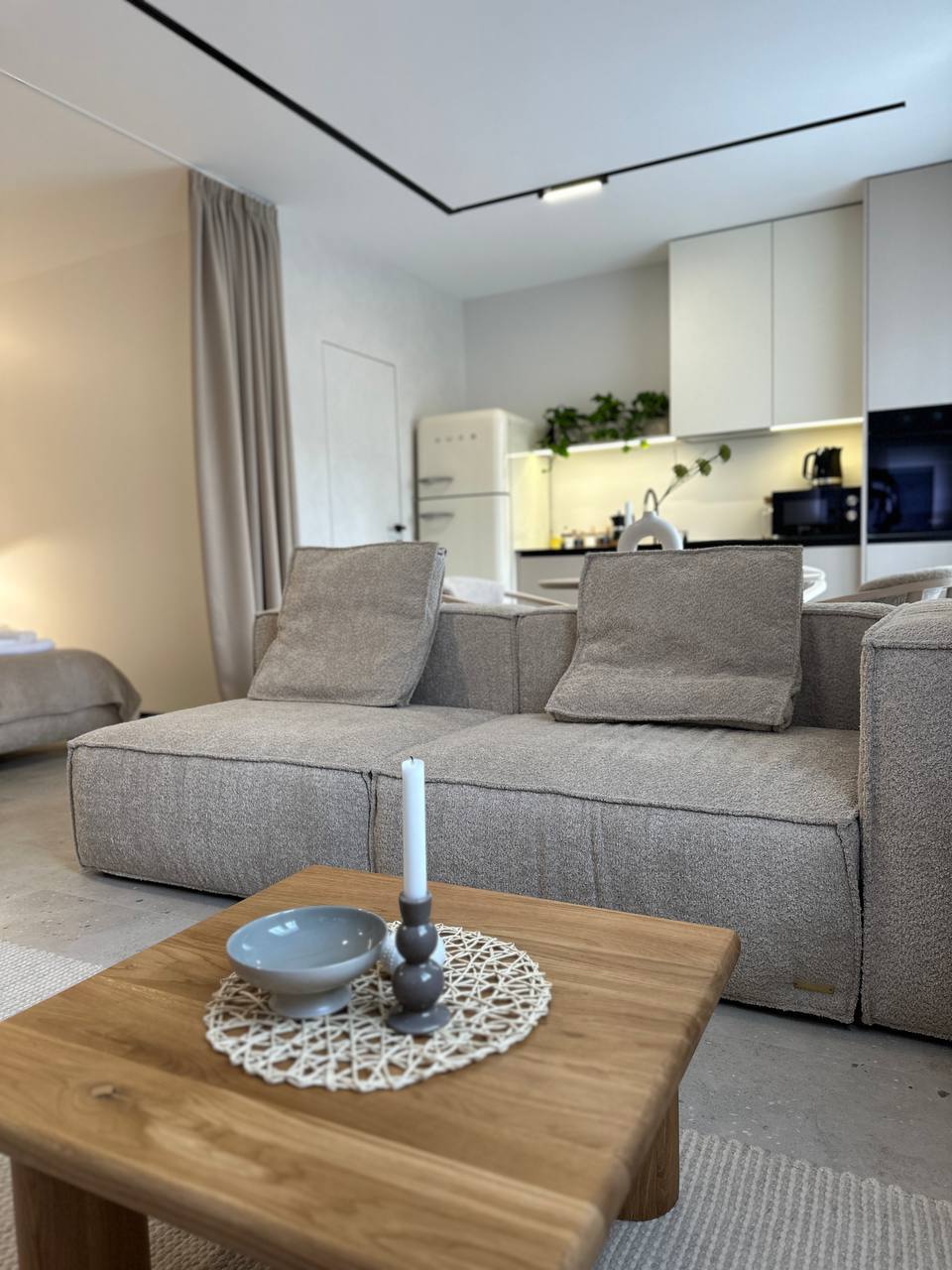 A comfortable living space is depicted, featuring a cozy sofa with two cushions. A wooden table displays a decorative bowl and a candle, while the kitchen area is visible in the background, showcasing modern appliances and greenery.
