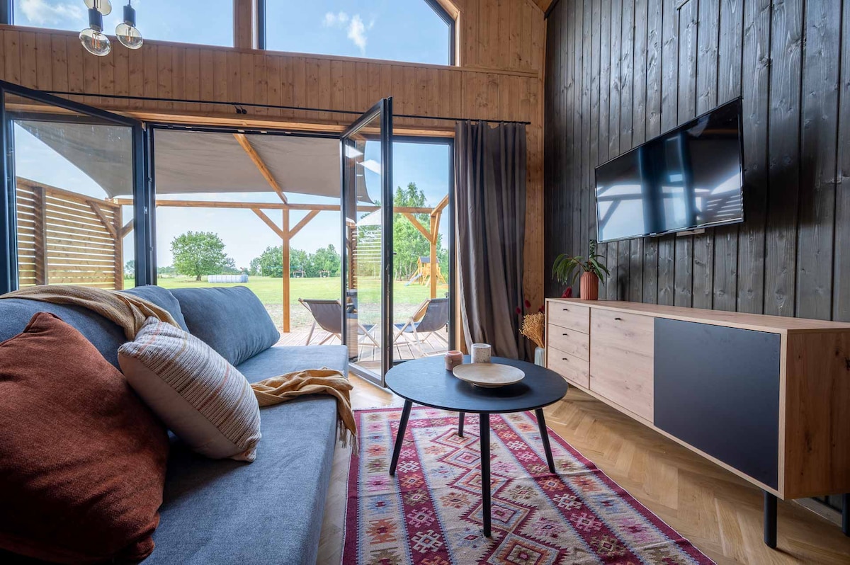A modern living area features a gray sofa adorned with colorful cushions and a warm throw. Natural light fills the space through large glass doors that open to a deck. A television is mounted on the wall above a sleek wooden media console, complemented by a round coffee table.