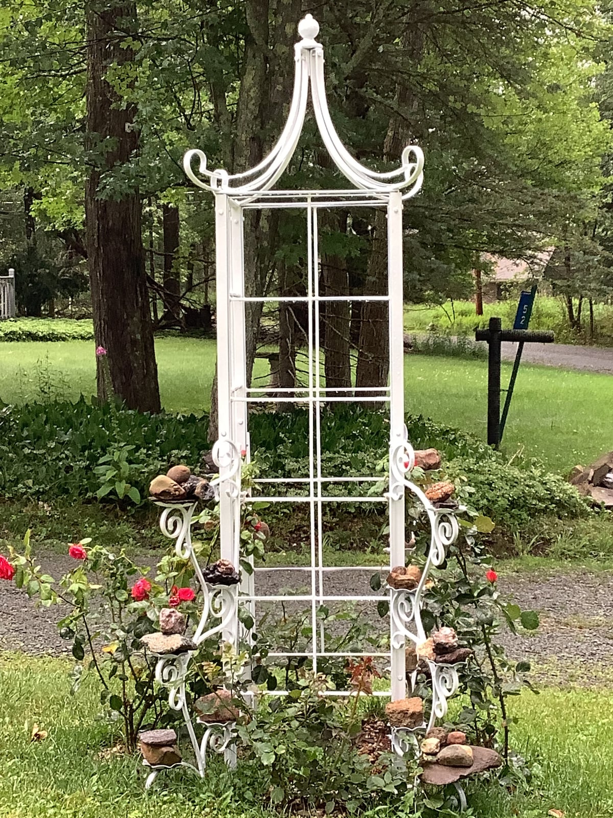 A white metal trellis stands in a lush green garden, surrounded by blooming roses and decorative rocks. The structure features an intricate design with multiple vertical and horizontal bars, providing a charming focal point amidst the greenery.