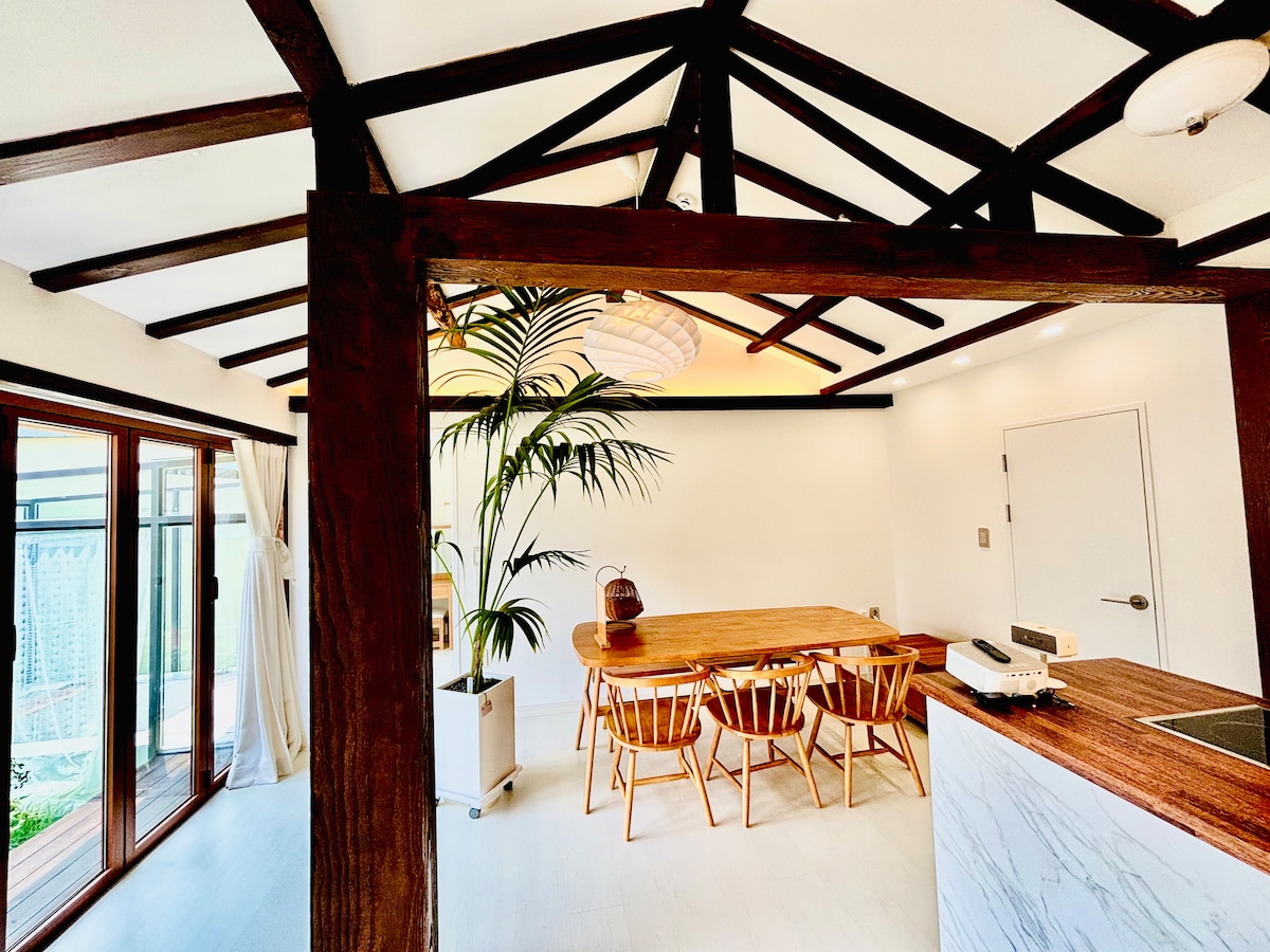 The dining room features a wooden table surrounded by six wooden chairs, with a large window allowing natural light to enter. A potted plant adds a touch of greenery, while a beamed ceiling enhances the room's spacious feel. A projector is positioned on the counter.