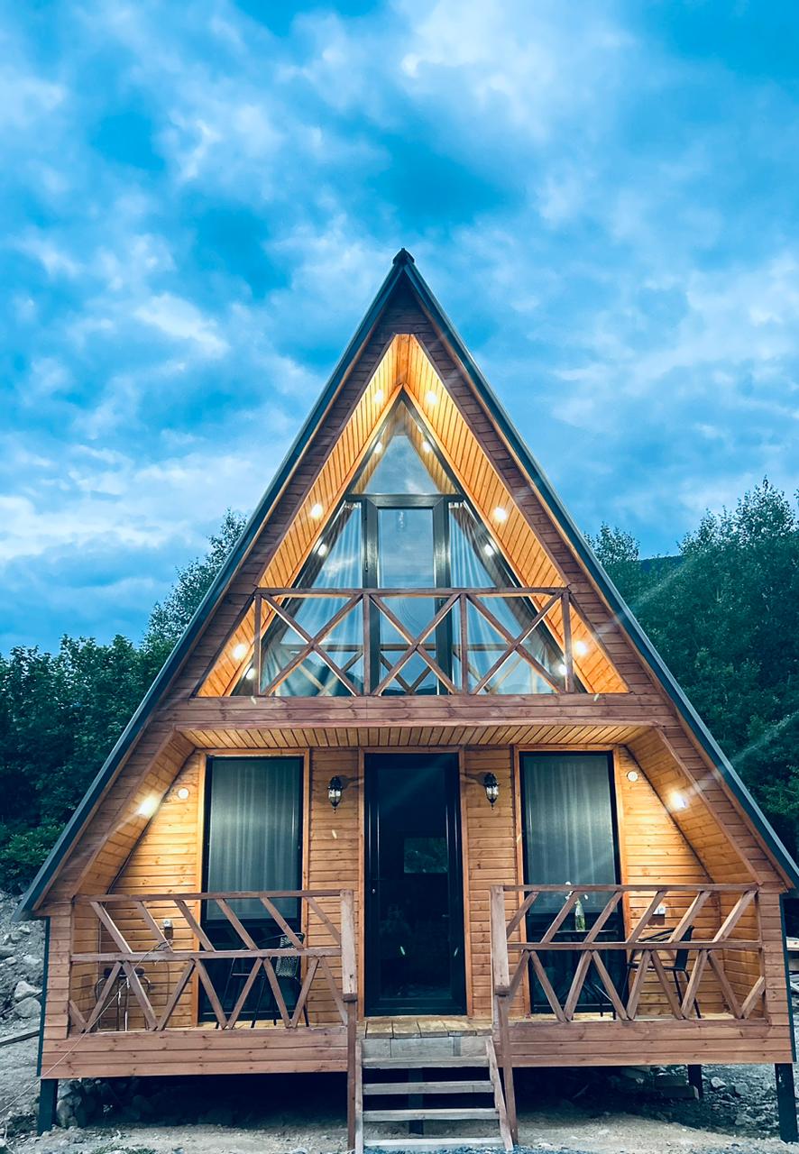 The exterior of a wooden A-frame house is illuminated under a twilight sky, featuring an expansive porch with railings. Large glass windows are framed by natural wood, providing a welcoming entry. The symmetrical roofline enhances the home's distinctive design.