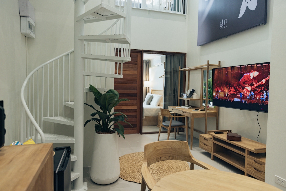 A modern living space is featured with a spiral staircase leading to the upper level. A comfortable seating area is arranged around a media console with a television. Natural light fills the room, highlighting a decorative plant in a white pot and wooden furniture.