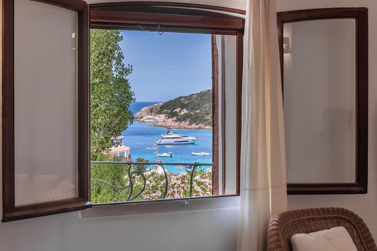 A scenic view from a window reveals the sparkling blue waters of the sea, framed by lush greenery and stunning rocky coastline. Boats can be seen gently floating on the water, enhancing the tranquil atmosphere of the surroundings.