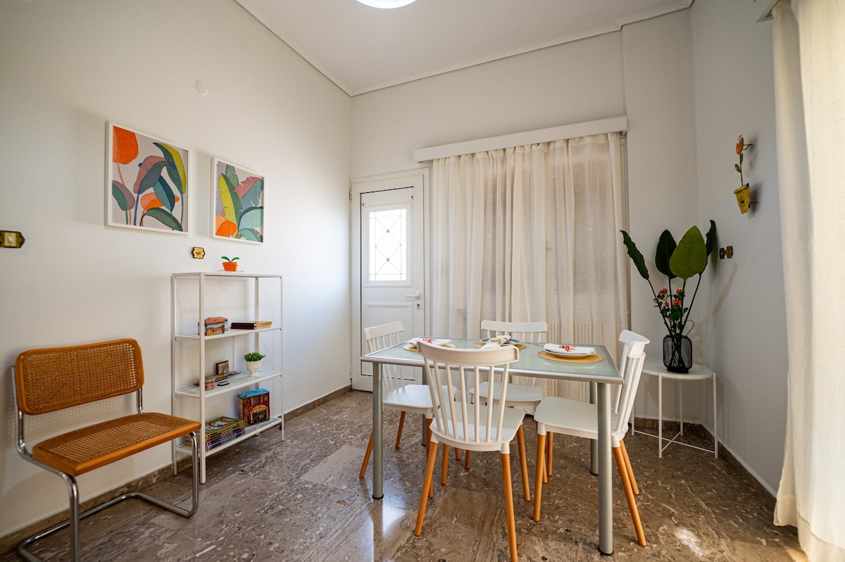 A bright dining area features a table set for four, surrounded by white chairs with wooden legs. Artwork adorns the walls, adding a splash of color. Natural light filters through sheer curtains, enhancing the open and airy feel of the space.