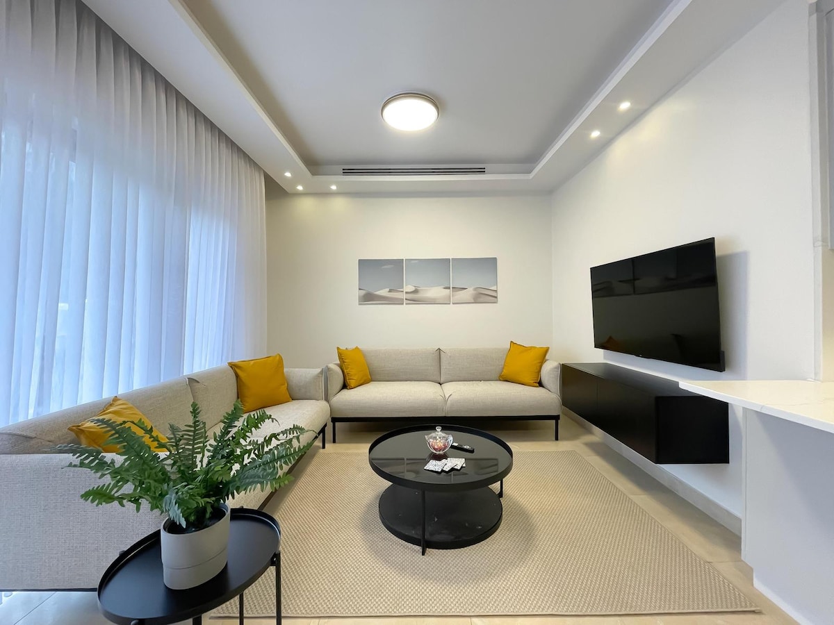 A modern living area is presented with a spacious, light-colored sectional sofa adorned with mustard yellow cushions. A round black coffee table sits in front, with decorative items placed on it. Natural light filters through sheer curtains, enhancing the inviting ambiance.