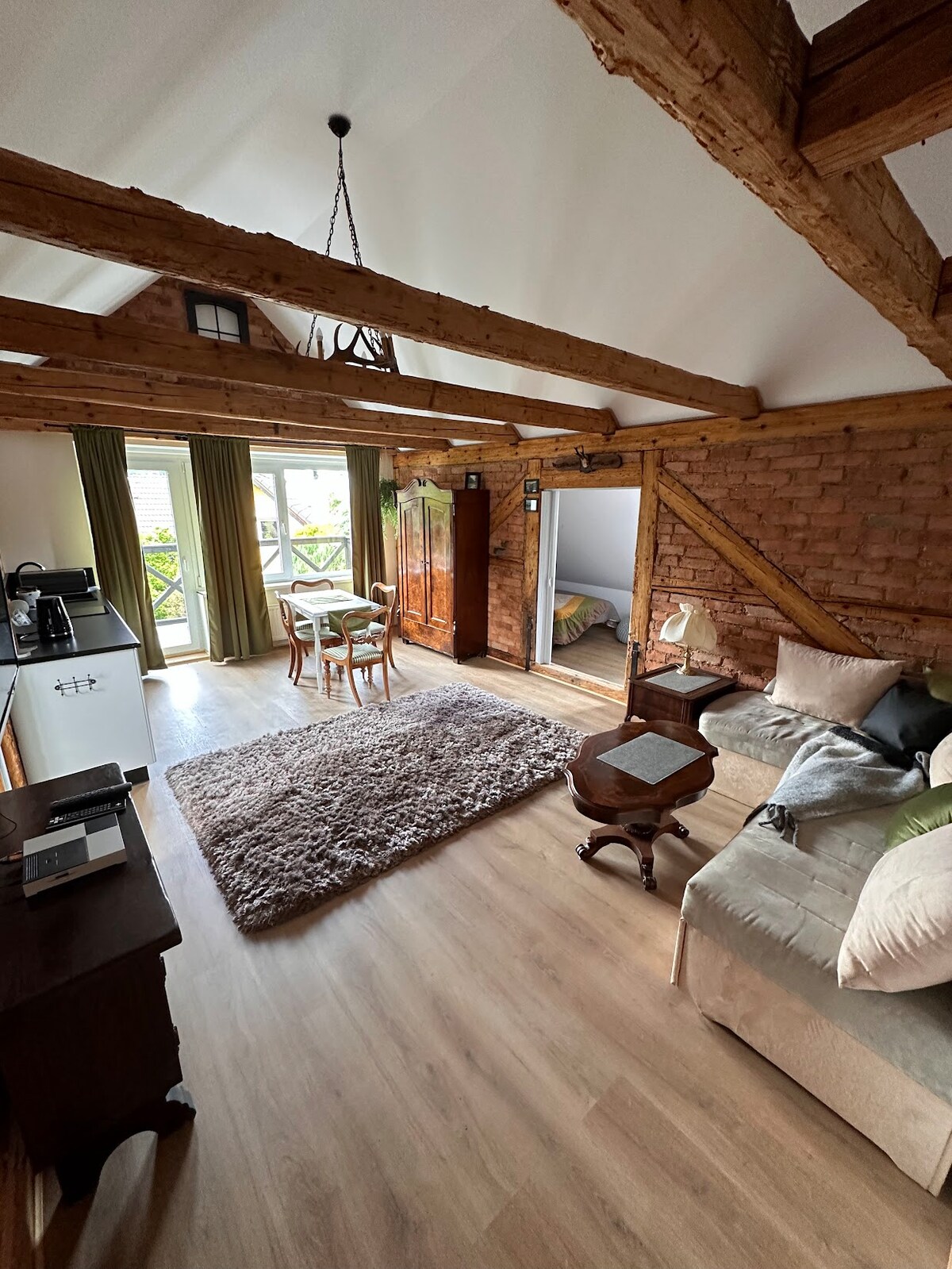 An open living area displays rustic wooden beams and a cozy seating arrangement featuring soft cushions. A dining table with chairs is positioned beside large windows, allowing natural light. A plush area rug is centered on the wooden floor, creating a warm atmosphere.