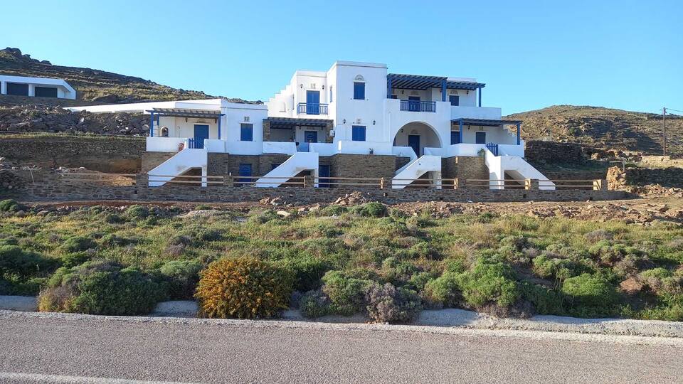 A modern two-story building is positioned against a clear blue sky, characterized by white walls and blue accents. Steep staircases lead to the entrance, surrounded by a natural landscape of low-growing greenery and rocky terrain.