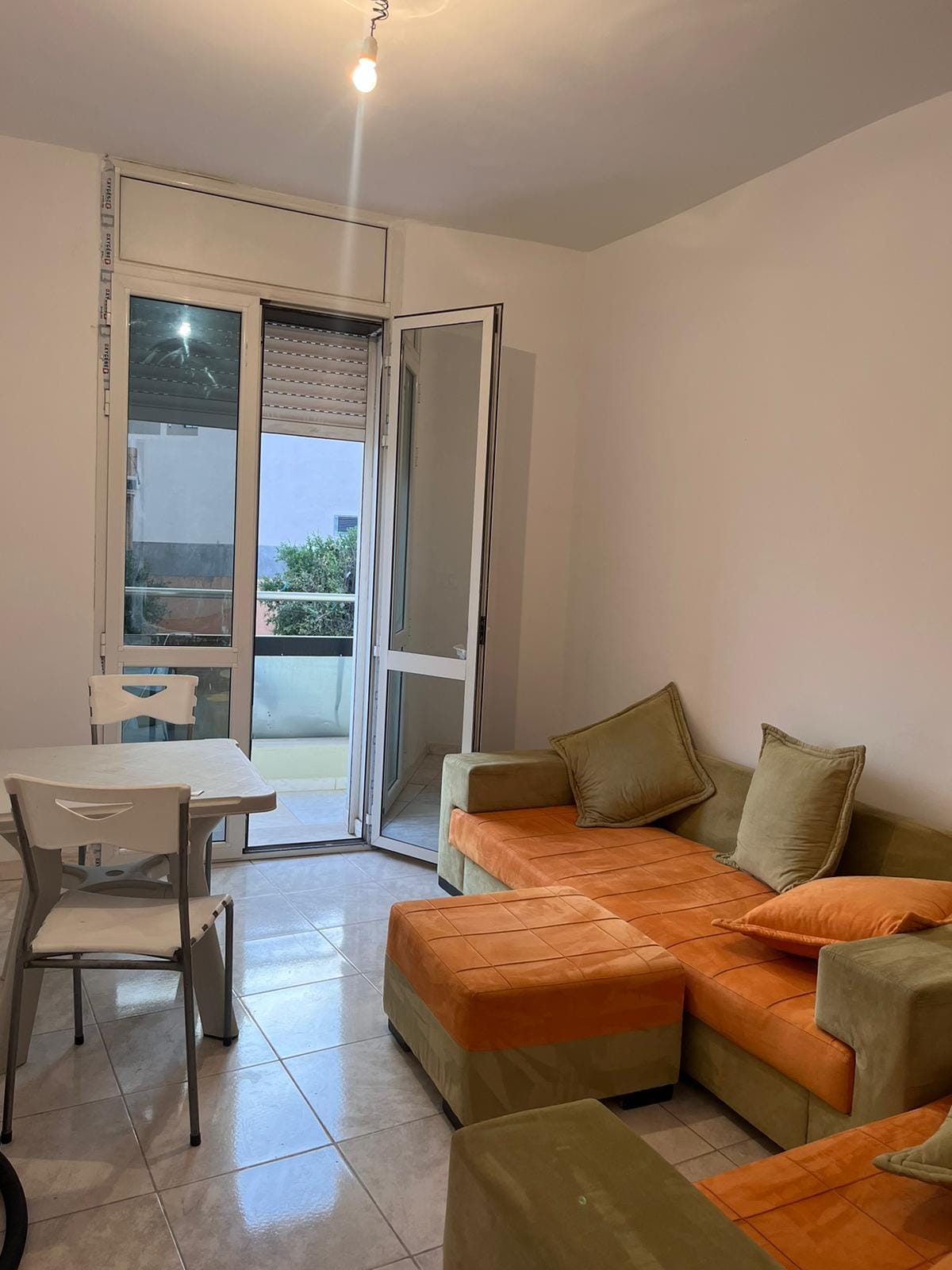 A bright living area features a corner sofa with orange cushions and a small dining table. Large glass doors provide access to a balcony, filling the room with natural light. Neutral wall tones and tiled flooring create a clean and airy space.