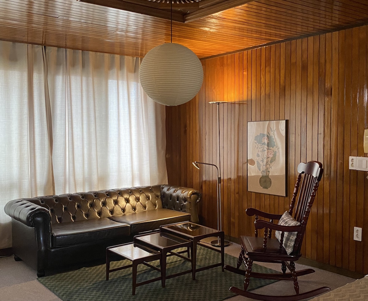 A cozy living area features dark wood paneling and a classic leather sofa. A rocking chair sits beside a small side table. Natural light filters through sheer curtains, illuminating a simple artwork displayed on the wall. A lamp provides additional lighting, creating a warm ambiance.