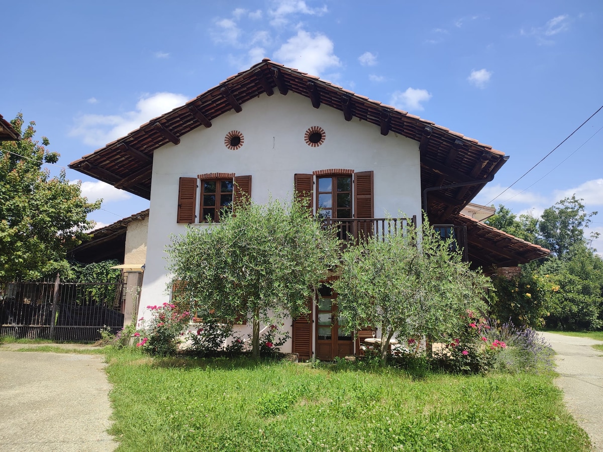 The exterior of the rustic house features a charming façade with white walls and wooden shutters. A healthy green lawn surrounds the home, complemented by small bushes and trees. A covered balcony extends from the upper level, inviting outdoor relaxation.