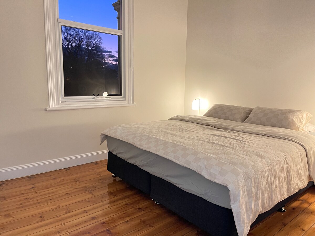 A spacious bedroom features a comfortable bed adorned with a light-colored quilt. A warm glow is emitted from a bedside lamp, and natural light filters in through a large window, which frames a view of the evening sky and trees.