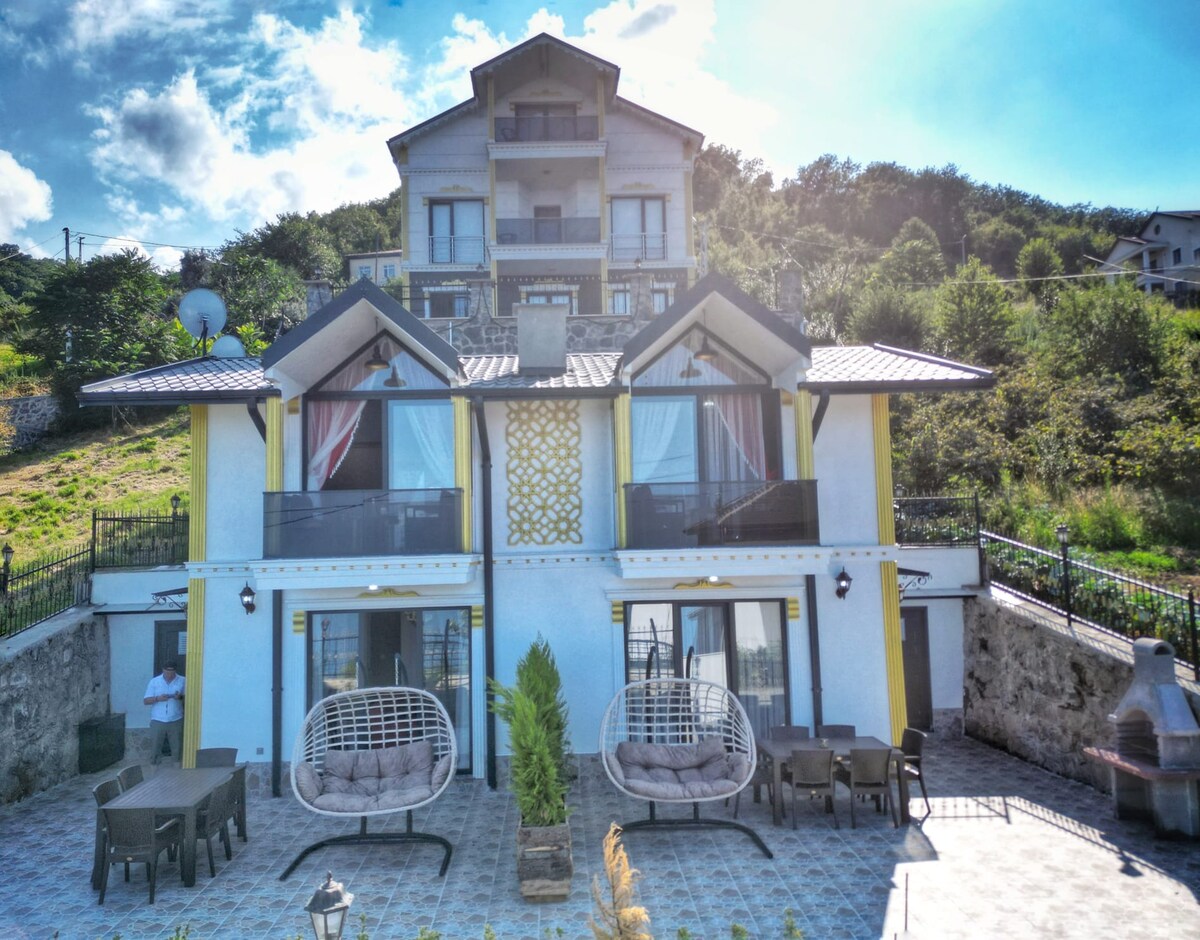 The villa showcases a two-story design with large glass doors leading to an outdoor patio. Two hanging chairs are positioned on the patio, surrounded by green landscaping. A barbecue area is visible, and the building features a neutral color palette under a bright sky.