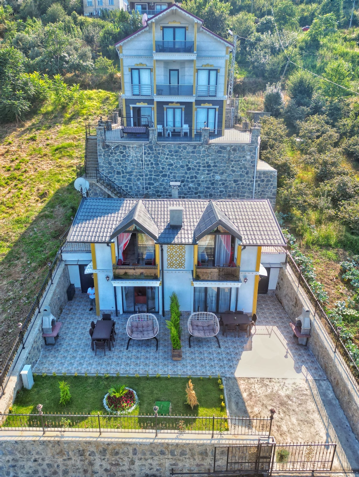 The image shows a multi-level villa surrounded by greenery. A spacious outdoor area features a manicured lawn with decorative plants, two hanging chairs, and a seating arrangement. The upper levels of the villa display multiple balconies, offering scenic views of the surrounding landscape.