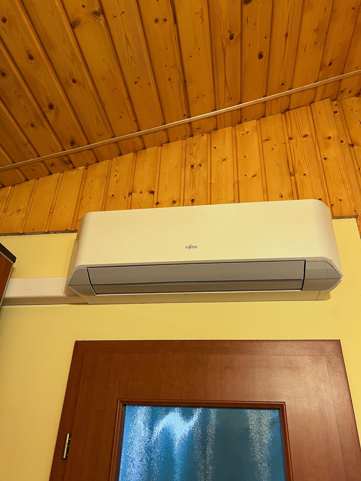 An air conditioning unit is mounted on a wooden ceiling, which features a natural finish. The wall below is painted in a calming yellow hue, while a wooden door with a glass panel is visible, adding to the functional design of the space.