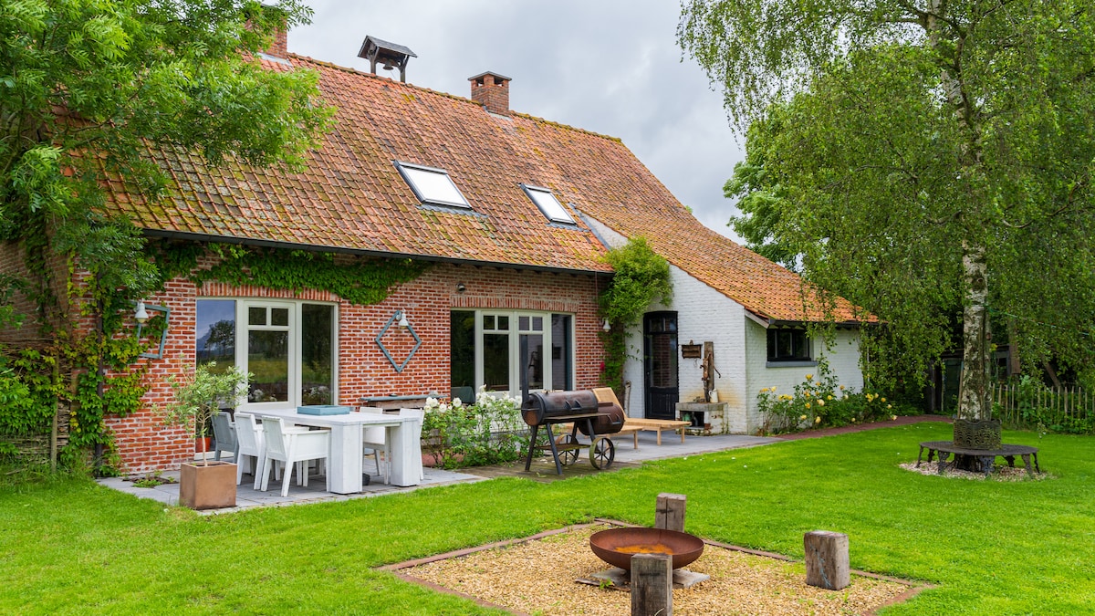 A charming cottage is surrounded by a vibrant green lawn. The exterior features a mixture of brick and white walls with multiple windows. In the foreground, a fire pit is situated on gravel, with outdoor seating arranged around it. A BBQ grill is visible nearby.