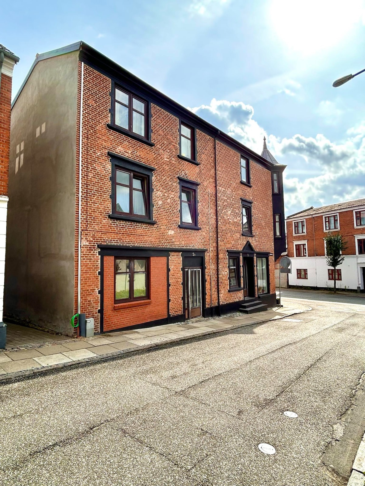A three-story brick building with a distinct façade featuring black window frames and door. The street is lined with a mix of buildings, and the scene is illuminated by natural light, suggesting a welcoming ambiance.