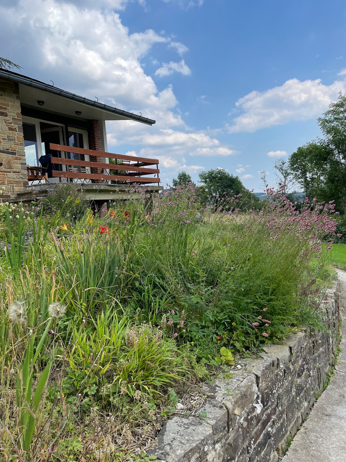 The exterior of the bungalow is shown, highlighting a landscaped yard with various greenery and blooming flowers. A wooden deck is visible above, providing access to indoor spaces. The sky is bright with scattered clouds, creating a serene backdrop.