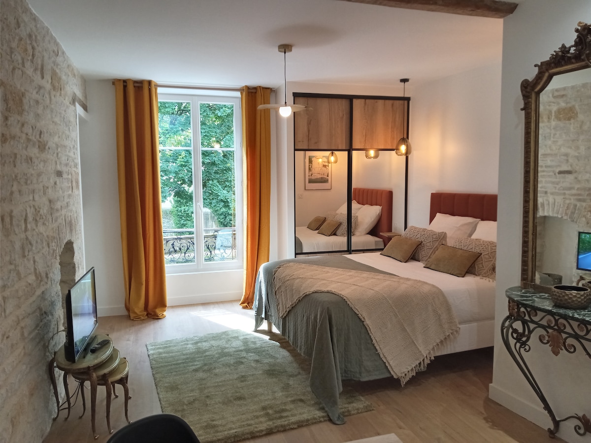 A well-lit sleeping area features a spacious bed adorned with decorative pillows and a neutral-toned blanket. Large windows with soft curtains allow natural light to fill the room, highlighting the textured stone wall and stylish mirror. A cozy rug completes the inviting space.