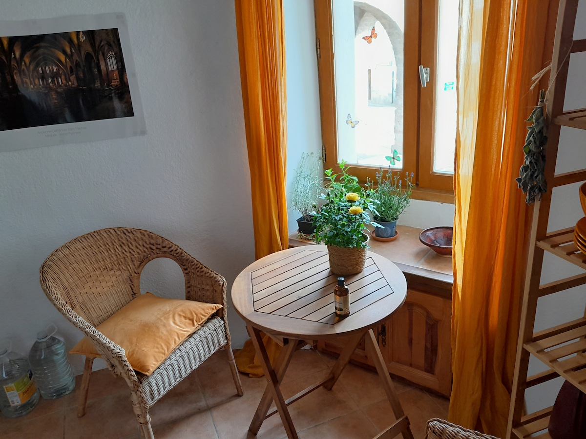 A cozy nook features a round wooden table and a wicker chair, illuminated by warm natural light. Yellow curtains frame the window, highlighting potted plants and a small decor item. Bottled water is placed nearby on the tiled floor, contributing to the inviting ambiance.