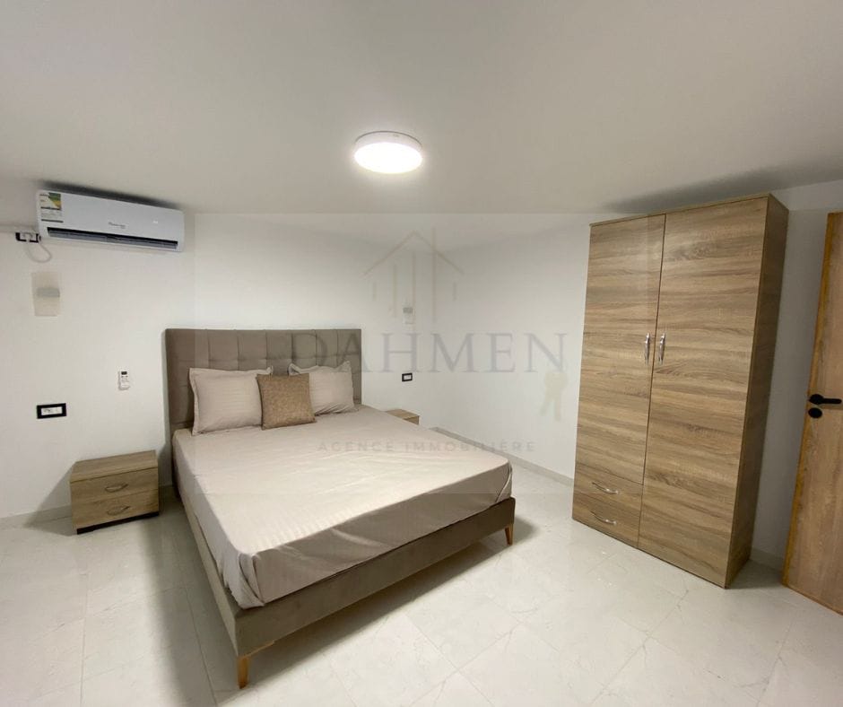 A spacious bedroom is presented, featuring a light-colored bed with a padded headboard. A minimalist nightstand and a wooden wardrobe are positioned nearby. Soft, neutral tones dominate the decor, while a ceiling light provides illumination. An air conditioning unit is discreetly mounted on the wall.