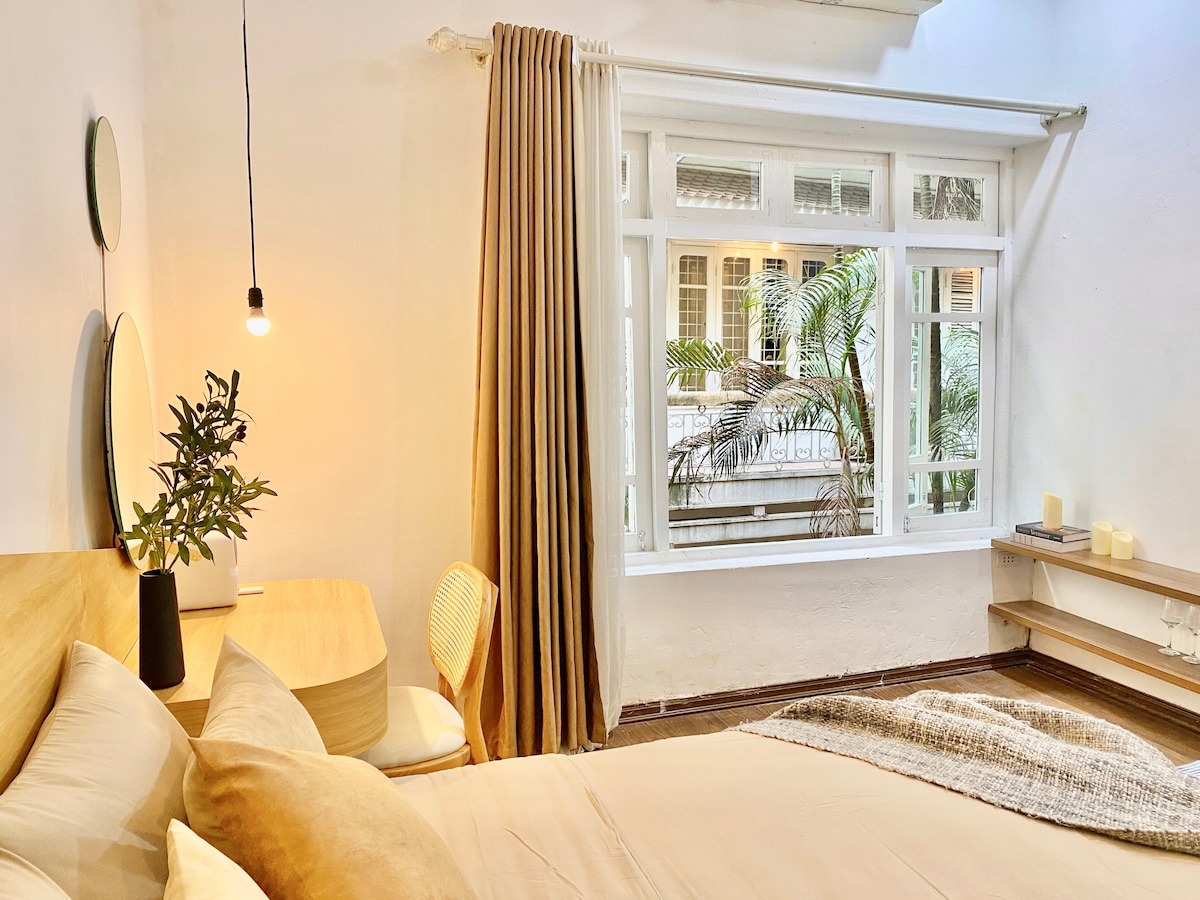 A spacious sleeping area is seen with a comfortable bed adorned in neutral tones. Natural light enters through a large window that overlooks lush greenery. A small bedside table and a modern chair complement the area, while curtains frame the window, creating a serene atmosphere.