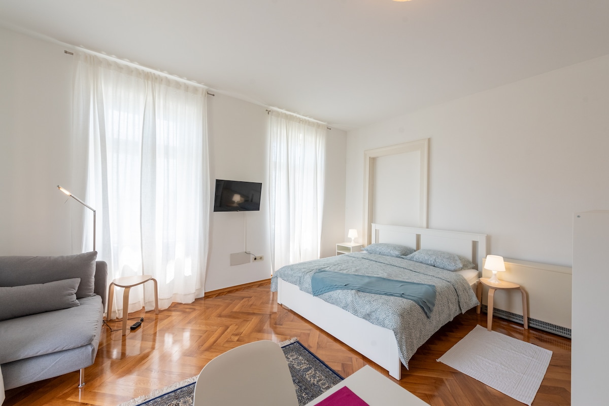 A bright room features large windows draped with sheer curtains, allowing natural light to illuminate the space. A spacious bed is adorned with light bedding. A comfortable sofa is positioned nearby, and a flat-screen TV is mounted on the wall adjacent to the seating area.