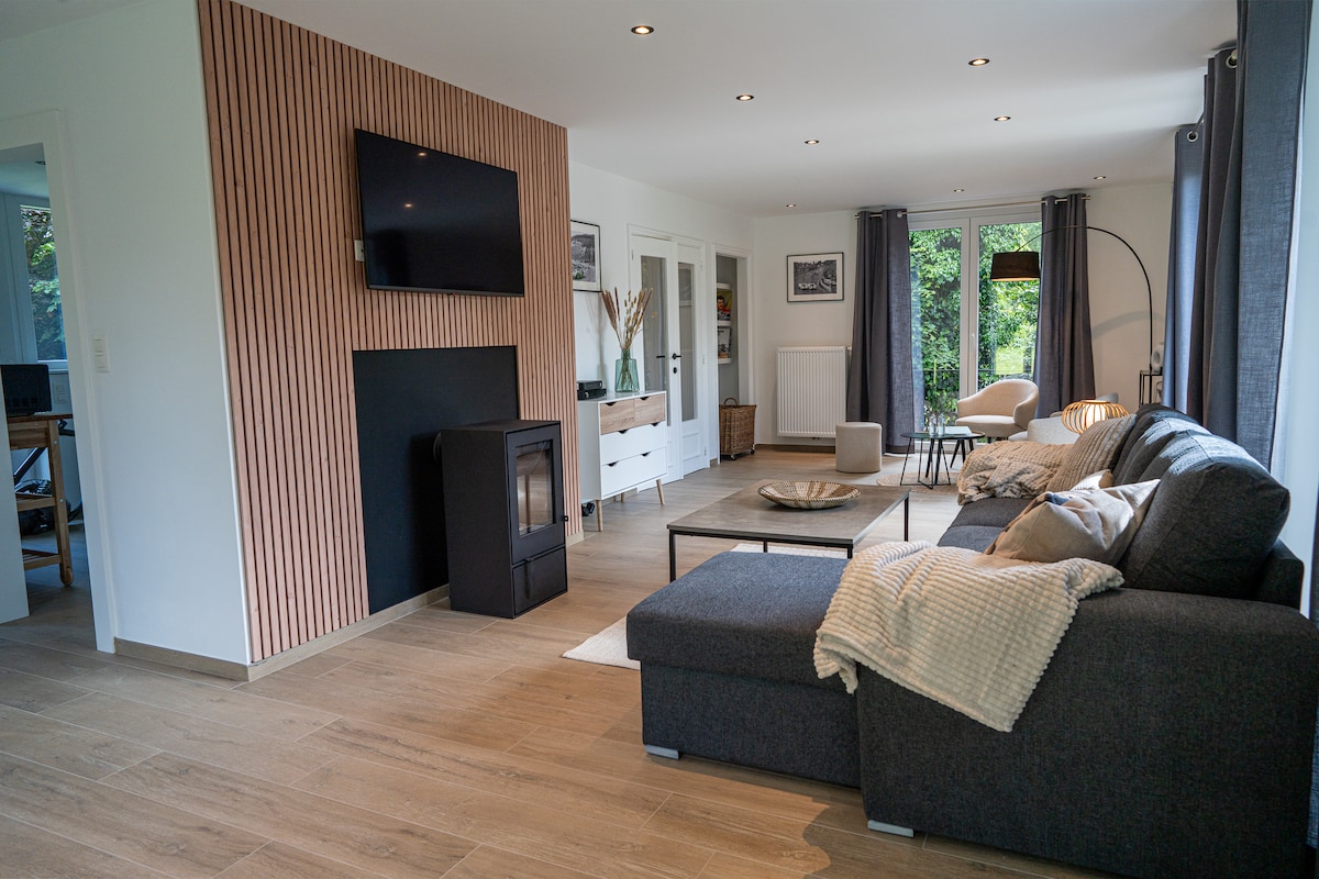 A spacious living area is featured, highlighted by a comfortable dark sofa positioned against a textured accent wall. A mounted television and an inviting fireplace add modern touches. Natural light filters through large windows, illuminating minimalist decor and cozy seating arrangements.