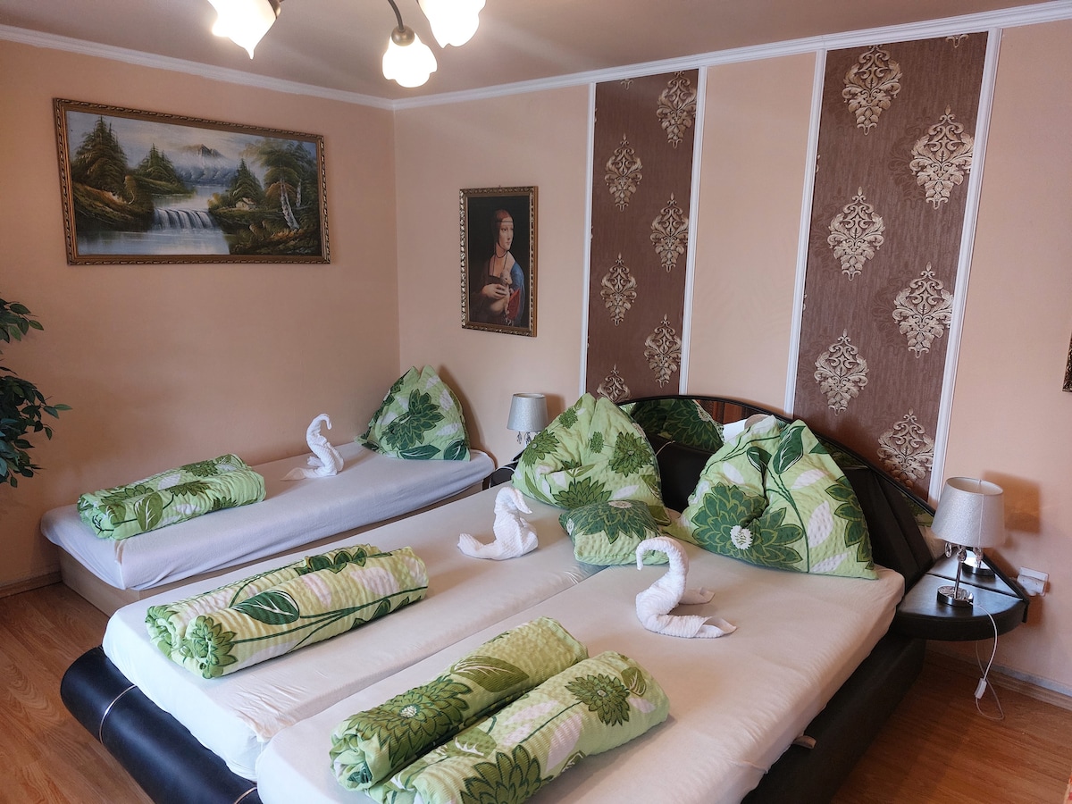 The room features two spacious beds adorned with patterned bed linens and decorative towels folded to resemble swans. Green floral accents on pillows create a relaxing atmosphere. Soft lighting is provided by matching bedside lamps, and a wall-mounted painting adds a touch of elegance.
