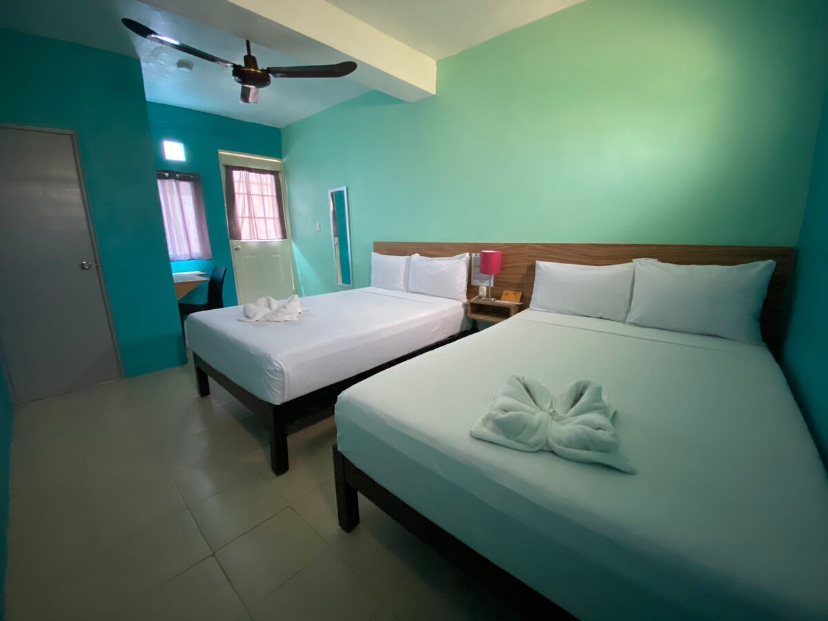 A bright and welcoming room features two spacious beds with neatly arranged linens. A ceiling fan provides comfort, while walls painted in soothing teal enhance the ambiance. A small table and chairs are positioned in an area near the window, allowing for natural light to fill the space.