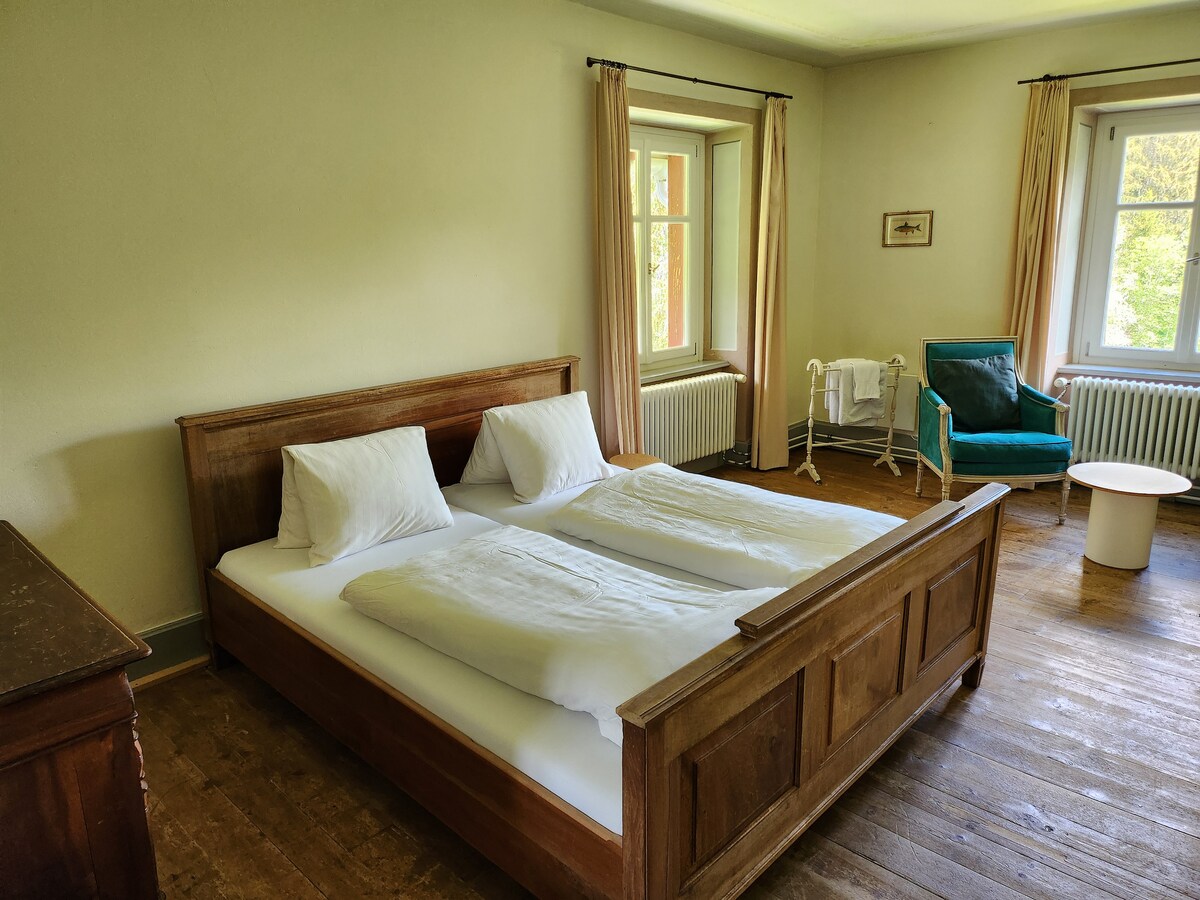 The spacious bedroom features a wooden bed with white linens and two pillows. Natural light fills the room through large windows, complemented by soft curtains. A cozy chair and a small table add to the simplicity, while a radiator is positioned beneath the window.