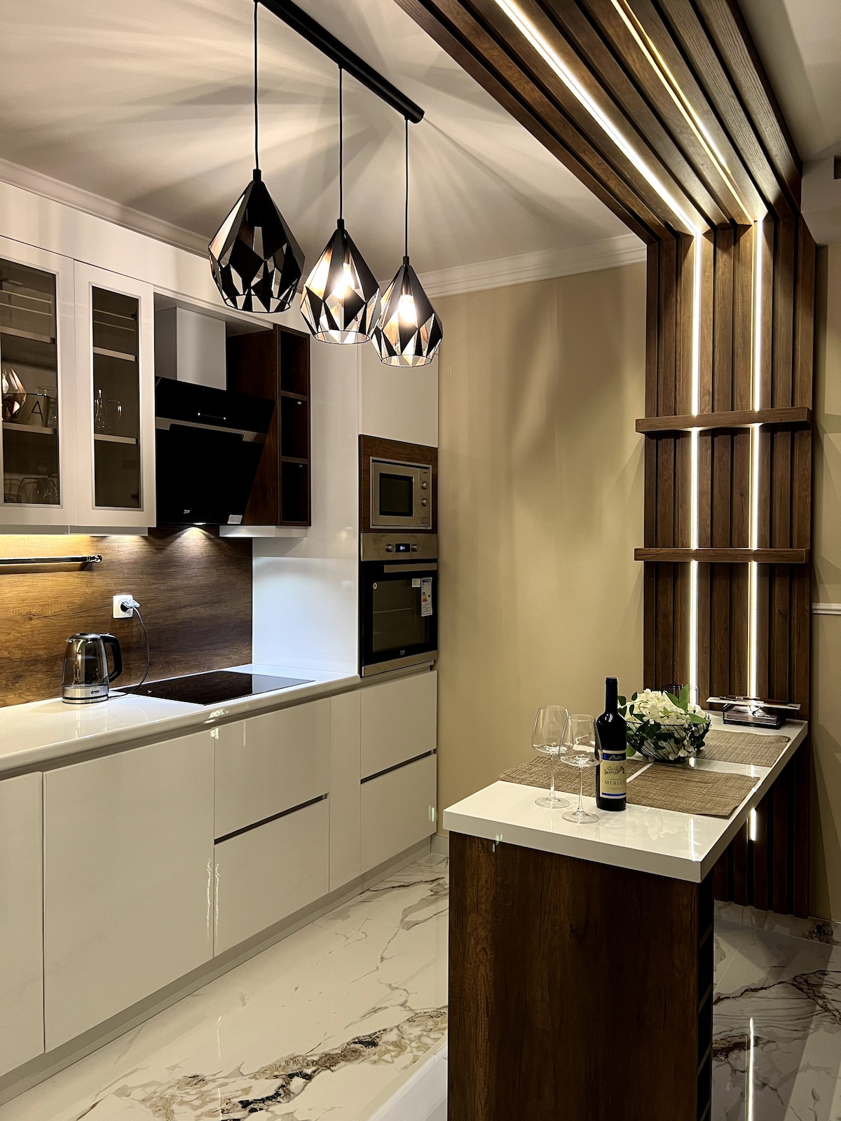 A modern kitchen features sleek white cabinetry and dark wood accents. Stylish pendant lights hang above a small dining area, accompanied by two glasses and a wine bottle. Modern appliances are integrated into the design, with light reflecting off the marble flooring.