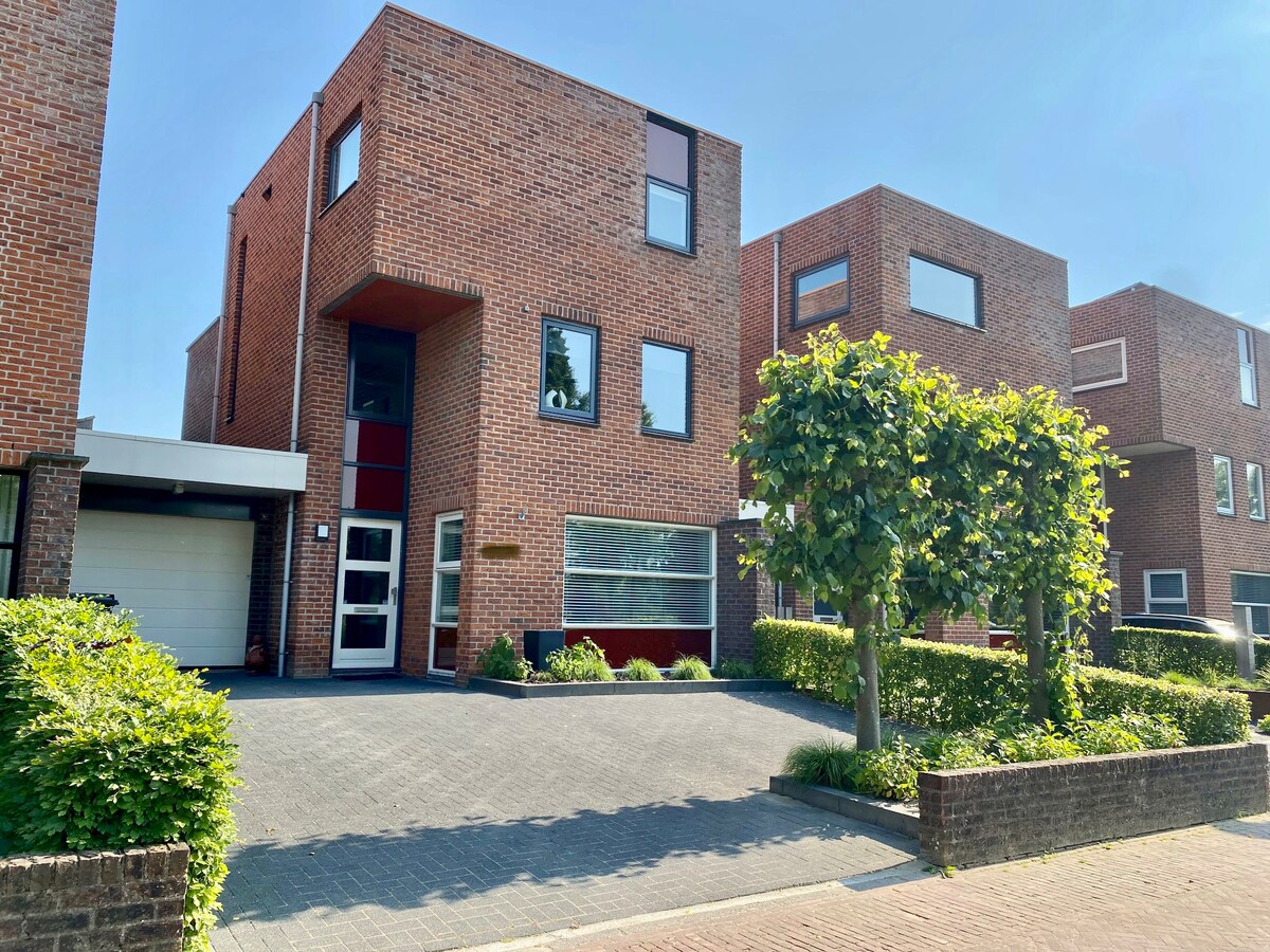 A modern, brick house with large windows is shown from the front. A paved driveway leads to the entrance, surrounded by neatly trimmed hedges and a small garden. Two small trees provide shade, while the blue sky enhances the clean lines of the architecture.