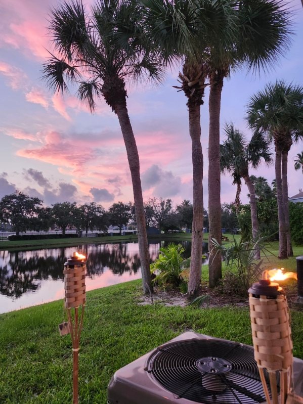 A serene outdoor scene captures the glowing reflections of a sunset on calm water. Framed by tall palm trees, tiki torches with flames add warmth. A small air conditioning unit is positioned in the foreground, emphasizing the lush greenery surrounding the picturesque view.