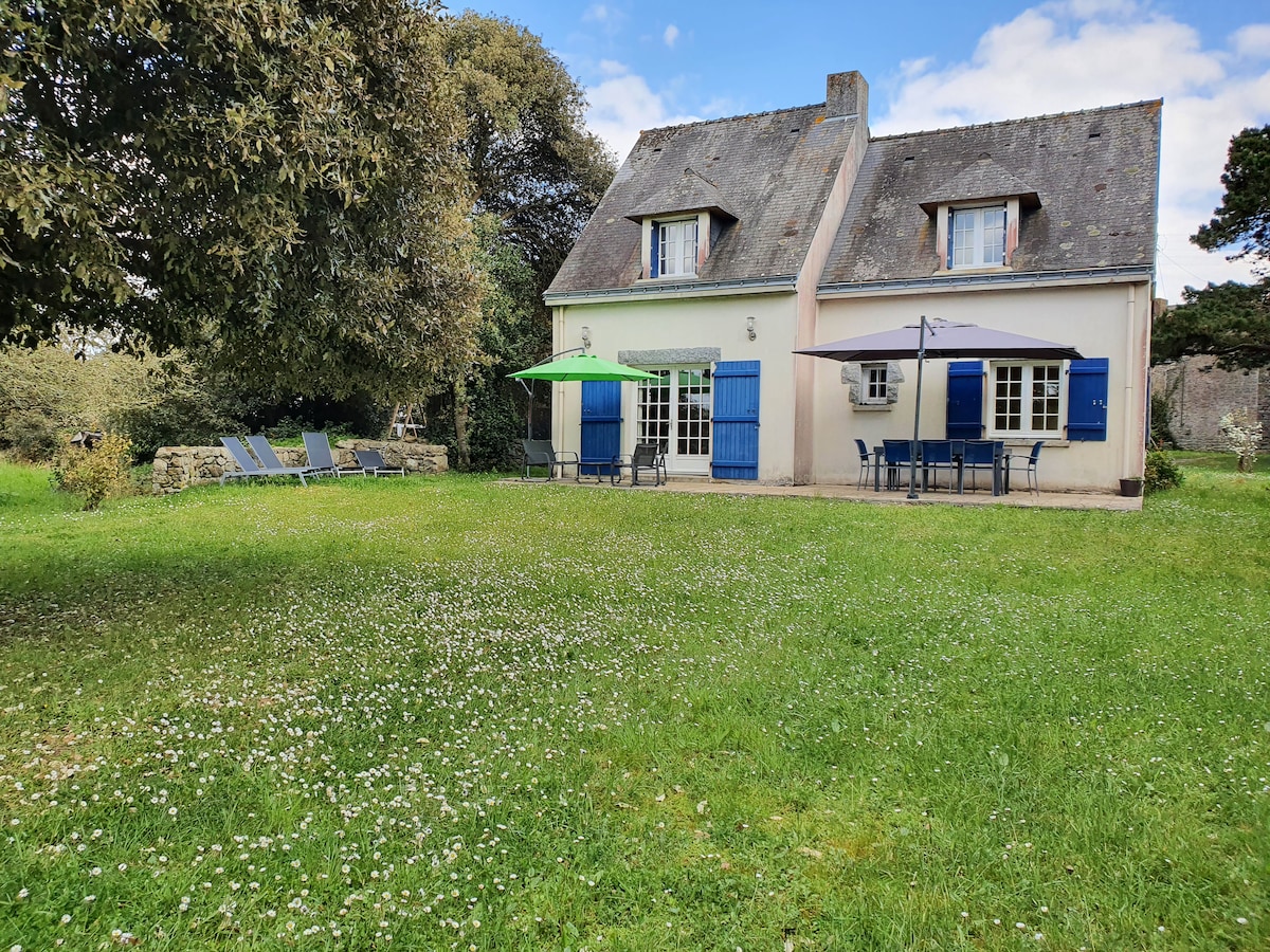 A charming house surrounded by a spacious green lawn features blue shutters and umbrellas. Sun loungers are positioned under trees, providing a relaxed outdoor space. The structure's design includes a gabled roof and multiple windows, enhancing natural light in the surroundings.