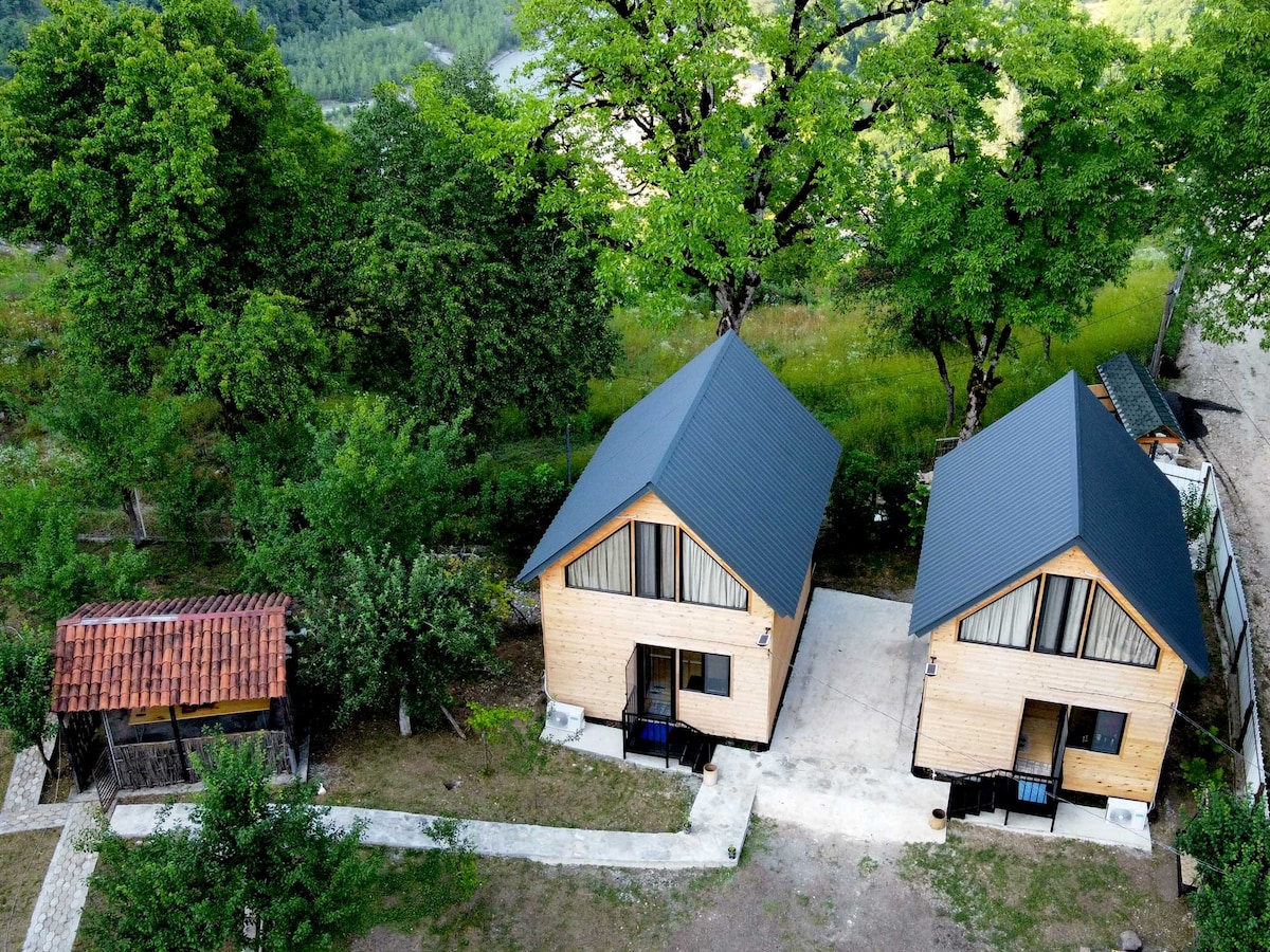 Two wooden cabins with sloped roofs are seen nestled in a natural landscape. A stone pathway connects the cabins and leads towards a small structure, while lush greenery surrounds the area, providing a serene backdrop.