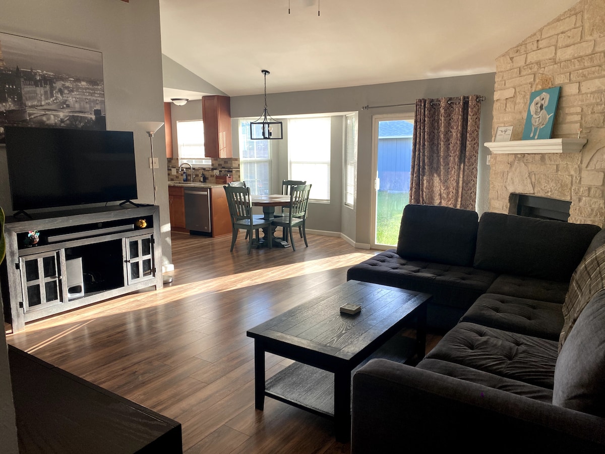 The open living area features a dark sectional sofa and a coffee table positioned in front of a large television. A dining table with green chairs is visible in the background, alongside a well-equipped kitchen. Natural light floods the space, highlighting the wooden flooring.