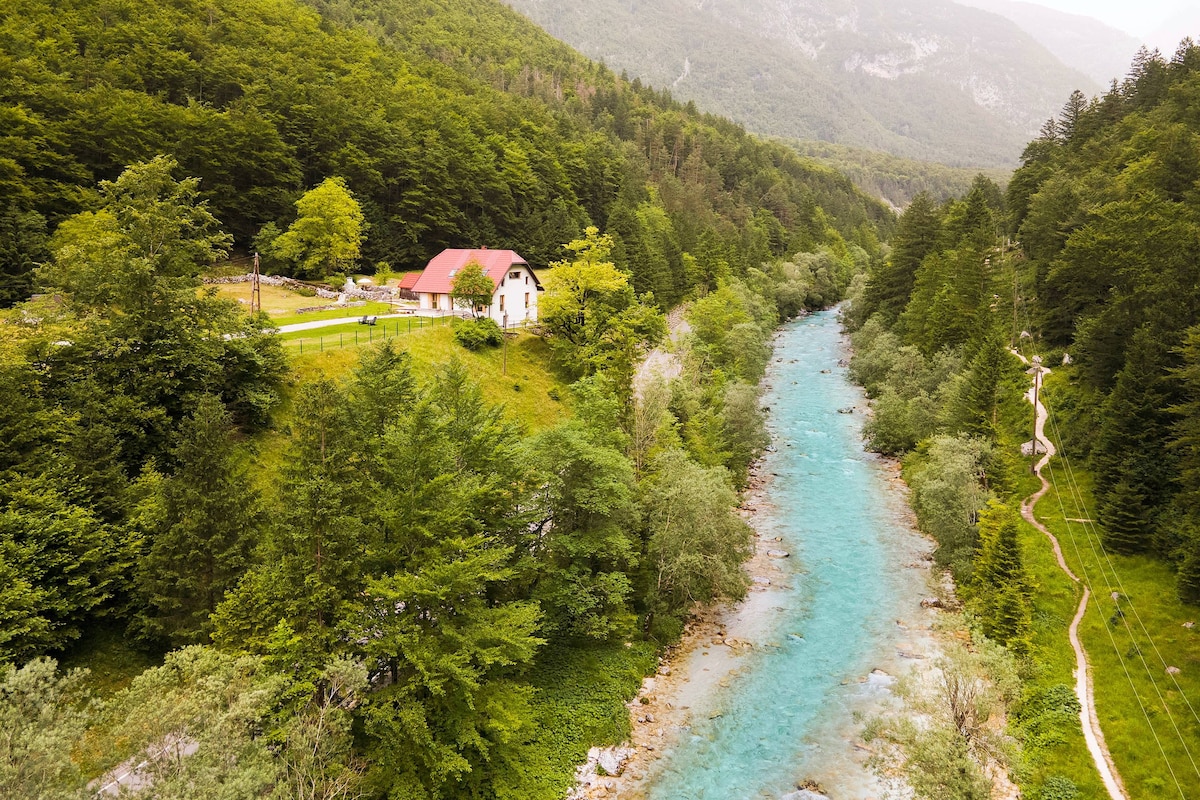 Aerial view of a cottage nestled among lush greenery along the banks of a turquoise river. Towering trees surround the property, while a walking path follows the river's edge, leading into the scenic landscape of rolling hills and distant mountains.