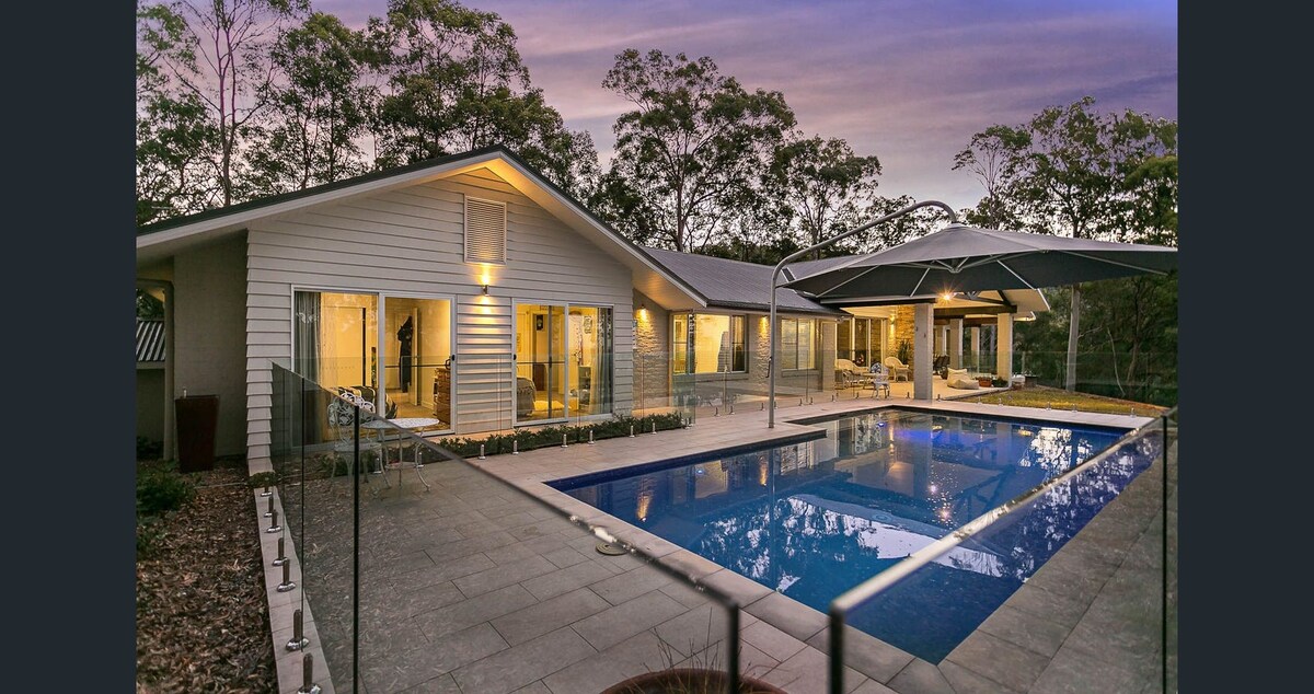 A modern home is showcased by the illuminated outdoor area, featuring a clear pool surrounded by spacious decks. Large glass doors provide views into the interior, blending indoor comforts with the natural landscape. Lush greenery serves as a serene backdrop.