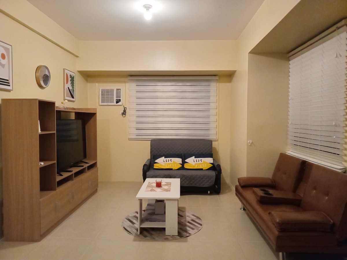 The living area features a cozy sofa and a pull-out bed, complemented by a compact coffee table in the center. A TV is mounted on the wooden media unit, and the space is illuminated by natural light filtering through the window with white blinds.
