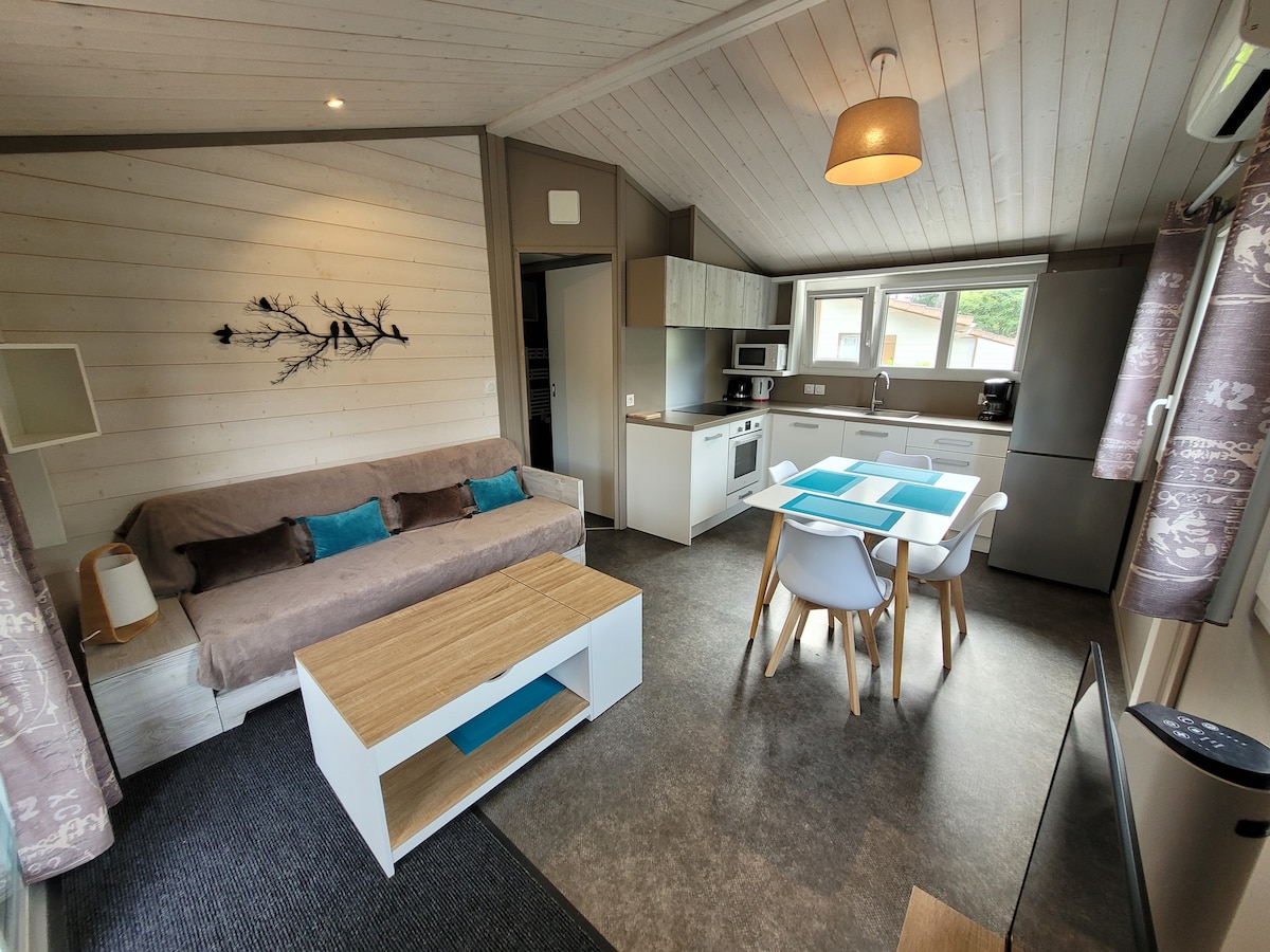 An open living area features a light-colored sofa with decorative cushions, paired with a modern dining table surrounded by sleek chairs. The kitchen area is equipped with essential appliances and cabinetry, while soft natural light enters through the windows dressed in patterned curtains.