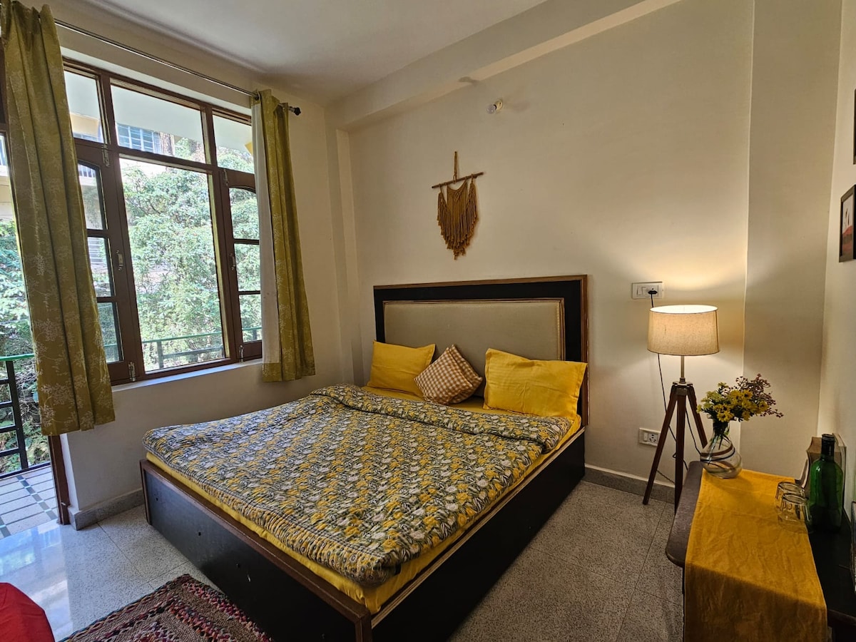 A cozy sleeping area is filled with natural light from large windows, adorned with leafy views. A spacious bed features bright yellow bedding and decorative pillows. A wooden side table holds a lamp, and a wall decoration adds subtle character.