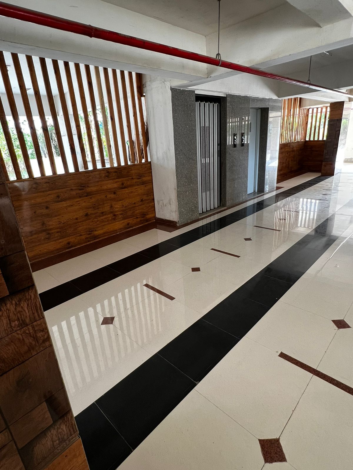 A spacious hallway is seen with a polished tile floor featuring a contrasting black and white pattern. Wooden accents enhance the welcoming ambiance, complemented by vertical wooden slats allowing natural light to filter through. Stone walls provide a modern touch to the contemporary design.