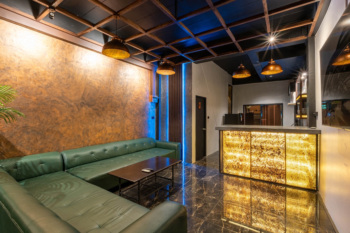 A spacious lounge area features a dark green sectional sofa arranged around a low coffee table. The walls exhibit a rich, textured finish, complemented by golden pendant lights above. A modern reception desk adds a touch of elegance, while dark marble flooring enhances the overall ambiance.