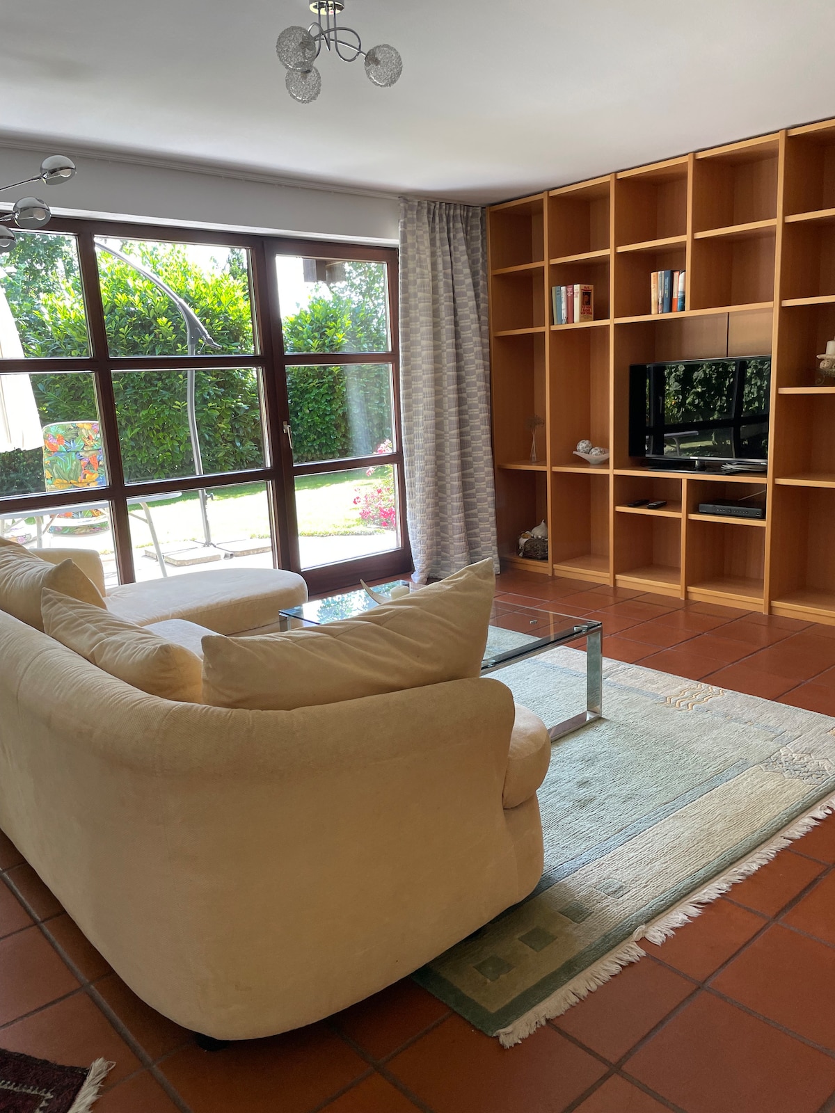 A sunlit living area features a plush light-colored sofa positioned near a glass coffee table. A built-in bookshelf lines the wall beside a large window that opens to a scenic garden view. Natural light enhances the inviting atmosphere of the space.