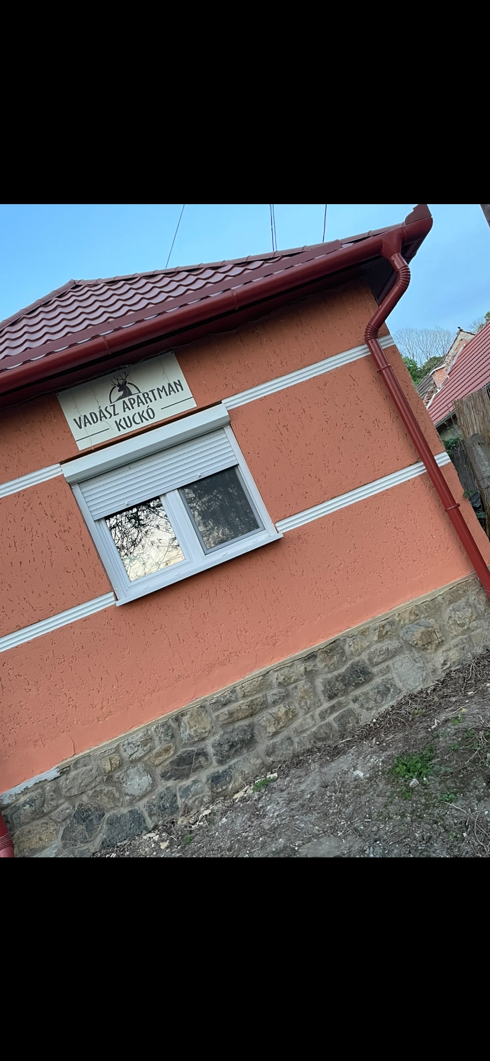 The exterior of the building showcases a warm orange facade with white decorative trim and a red roof. A rectangular window is centered on the wall, which is accented by stone detailing at the base. A sign is mounted above the window, identifying the location.