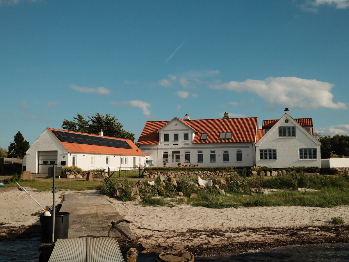 A spacious and well-maintained property is showcased, featuring a classic white exterior with red roof tiles. The surrounding area is landscaped with grassy patches and stone borders, and a serene waterfront is visible in the foreground.