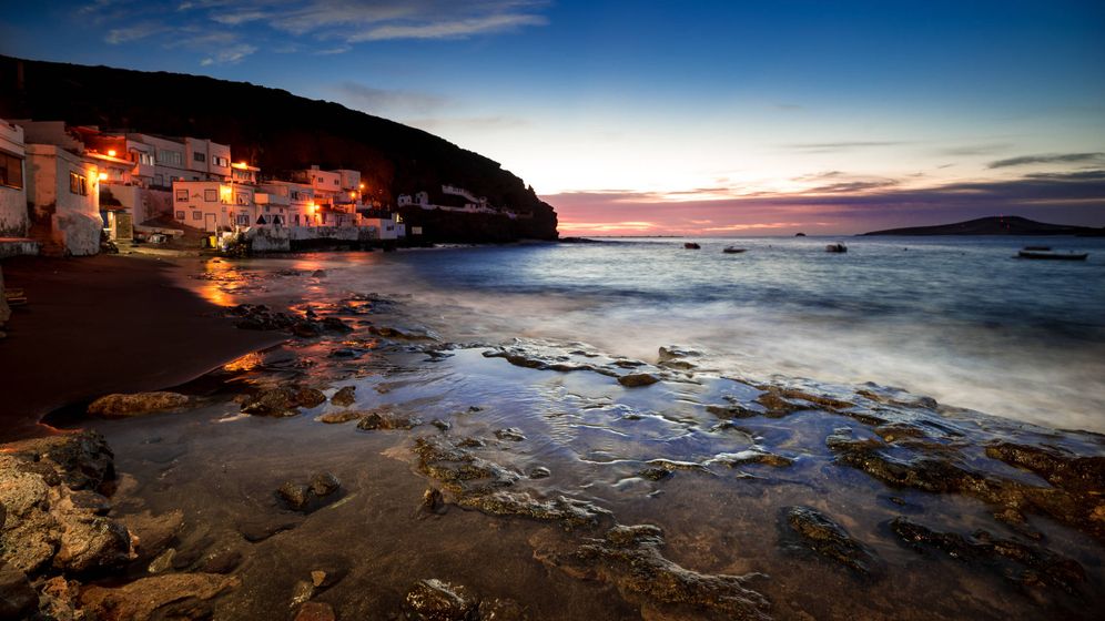 An ocean view at dusk captures the gentle waves lapping against a rocky shoreline. Soft orange lights illuminate nearby houses, creating a serene ambiance. The horizon displays subtle hues of blue and purple as the sun sets, offering a tranquil scene by the water's edge.