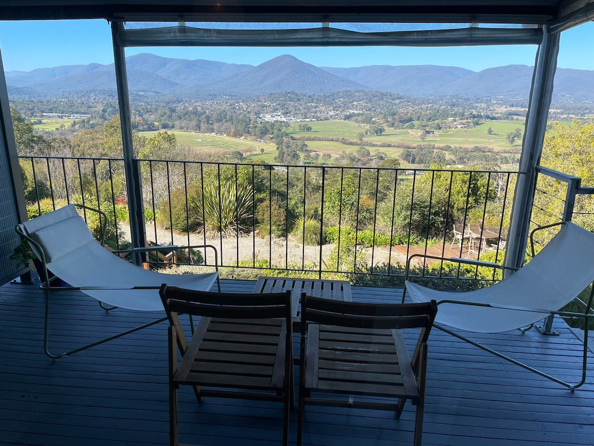 A spacious balcony features two lounge chairs and a wooden table, positioned to overlook expansive mountain views. The landscape showcases rolling hills and greenery, framed by a protective railing. Natural light enhances the serene atmosphere through the open sliding doors.
