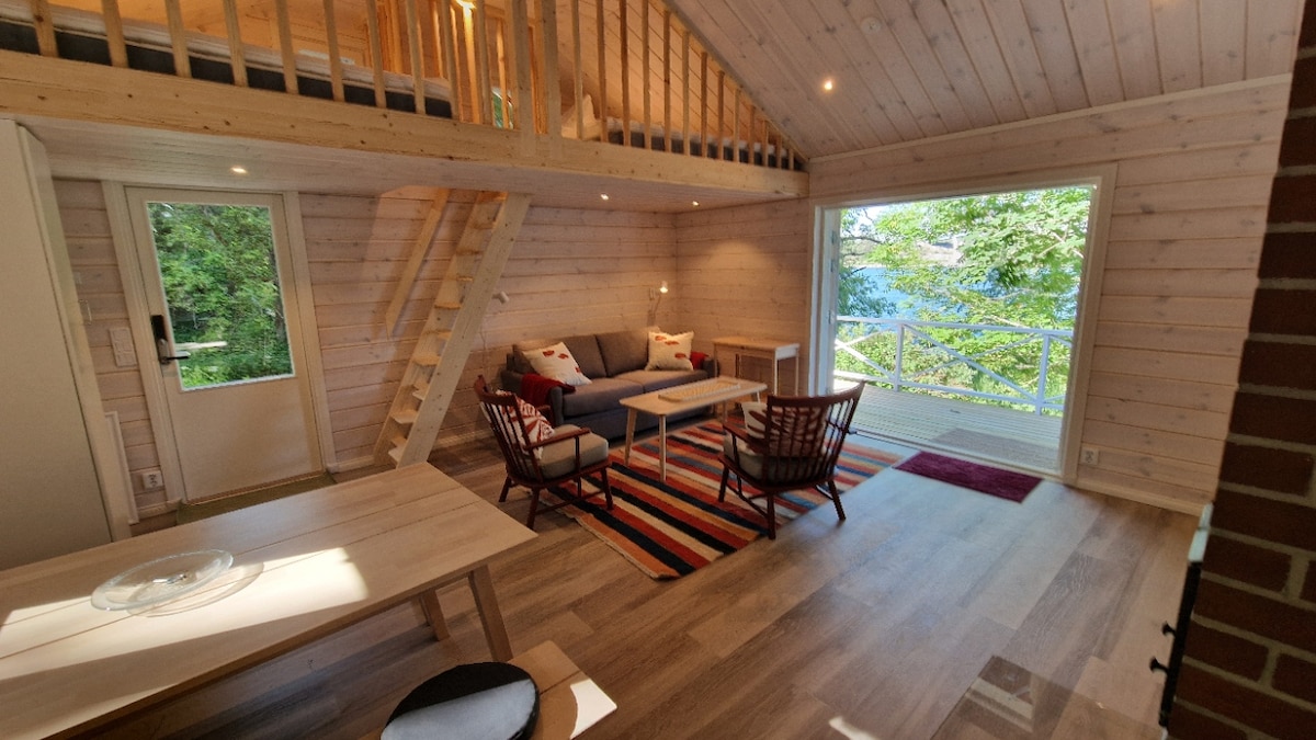 An open-plan living area is showcased with wood-paneled walls and large windows revealing the surrounding greenery. A comfortable sofa is paired with two accent chairs on a striped rug, while a dining table is positioned nearby. Natural light fills the room, enhancing its inviting ambiance.