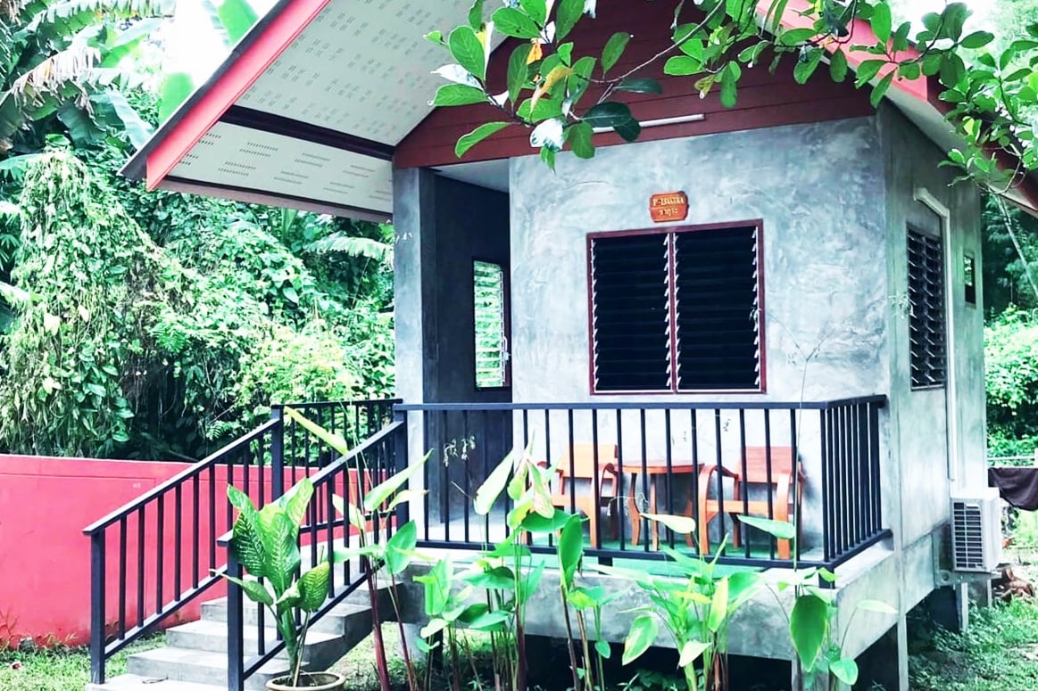 A small, unique house is surrounded by greenery, featuring a red roof and concrete walls. A porch wraps around one side, offering outdoor seating. Large windows provide views of the natural surroundings, while lush plants are situated at the base of the structure.
